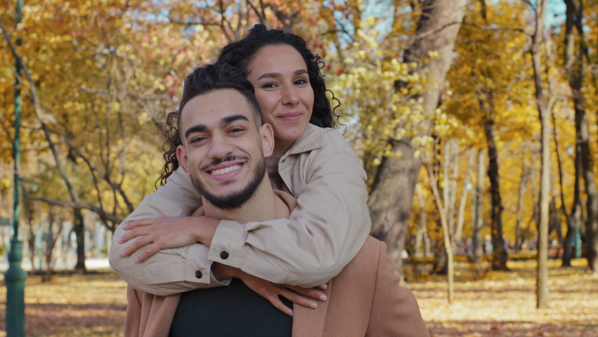 Close up hispanic couple on date in autumn park happily smiling attractive girl hug male by shoulders handsome bearded guy move holding piggyback female outdoors pair look up enjoying fall sunny day | Shutterstock HD Video #1083048556
