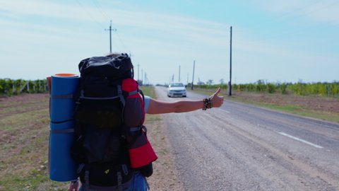 Woman with large tourist backpack, mat goes along road trying to stop car with raised hand gesture. Female hitchhiker, backpacker, hiker on roadside near field. Summer, afternoon. Back view