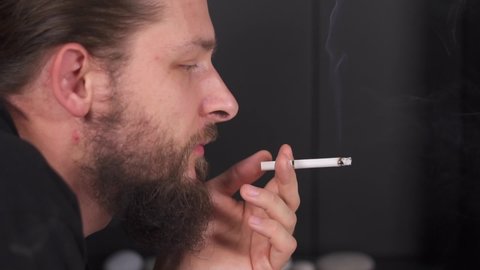 man smoke cigarette. Caucasian guy with beard look aside from side and blows smoke. Smoking is bad for your health. Light cigarette and light cigarette with a lighter. fire burns near person face.