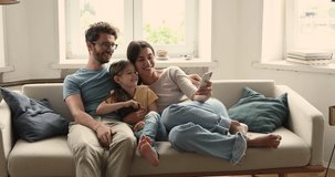 Friendly young mom dad small girl cuddle rest on comfy couch engaged in shopping online on cell watch photo video at social media. Happy family parents little child have fun with phone at home use app