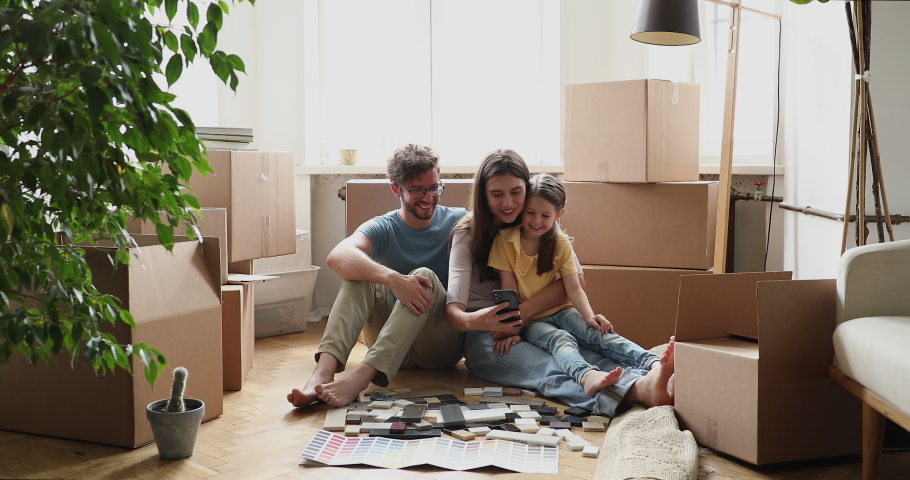 Happy spouses with preschool age kid girl make renovation at living room after moving in. Young female sit on floor hold phone show husband small daughter decor sample at web shop offer design variant Royalty-Free Stock Footage #1083055366
