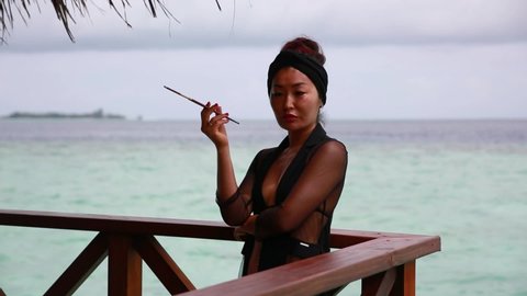 a girl in a black evening dress in a retro style stands at a water villa and smokes a cigarette through a mouthpiece against a background of blue sky and azure sea. Maldives resort.