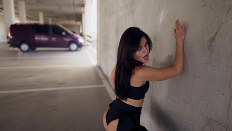 Dancing sexy woman performs modern dance, posing, twerking in parking zone leaning on wall