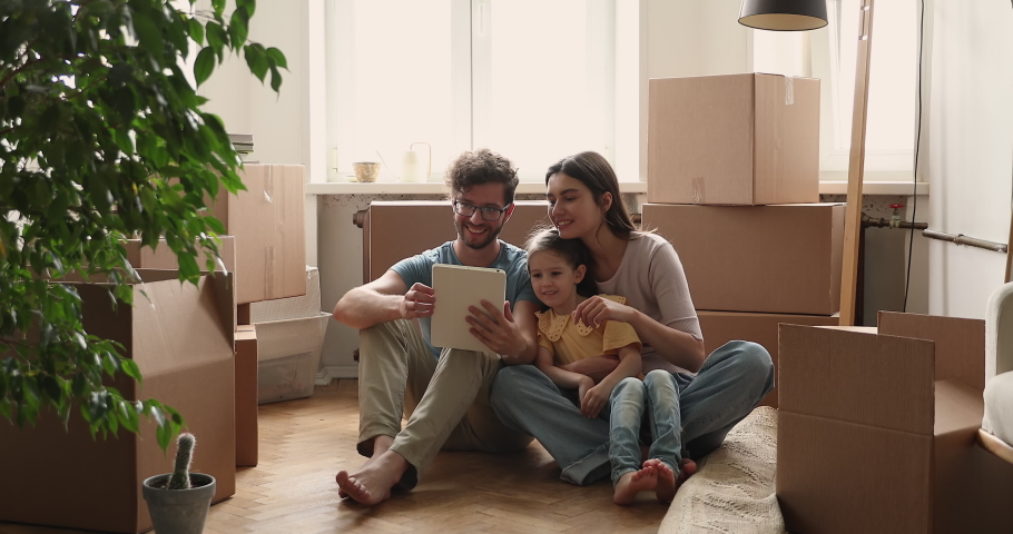 Excited young couple with child realty buyers sit on floor among carton boxes shoot funny selfie on tablet at new home. Mom dad little daughter do living room renovation choose design online using pad Royalty-Free Stock Footage #1083058141