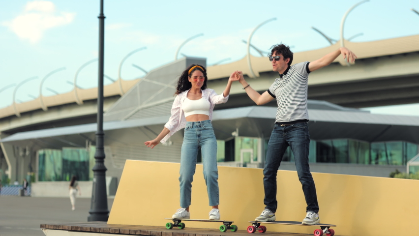 Active boyfriend and girlfriend learn to ride longboard together holding hands. Playful young couple laugh stand on skateboard. Man and woman wearing street style clothes skaters outdoor in urban area