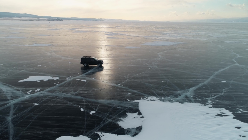 Aerial follow lonely car drives along endless cracked ice field of frozen winter lake Baikal at an orange sunset. Vehicle travel by car through untouched places Russia Siberia. Open space. 4k Royalty-Free Stock Footage #1083063853