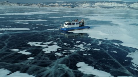 Flight backward above Tourist ship hovercraft fast rides on frozen blue ice winter lake Baikal khivus. Cracked ice field, open space. Mountains nature epic landscape. Trip travel Siberia Russia. Stock