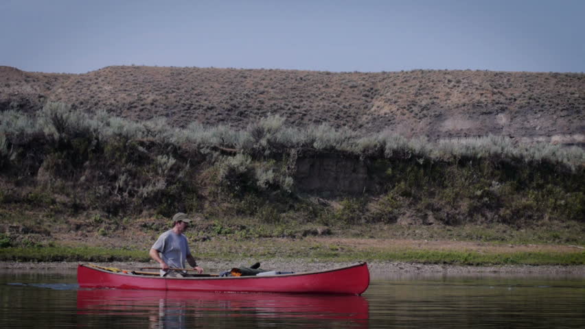 Man with a mustache fishes in a red canoe in a river with dolly like movement