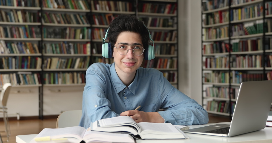 Student guy in eyeglasses and headphones sit at table with laptop and heap of textbooks, studying alone in college library, smile looking at camera. Education, e-learning, gen Z, modern tech concept Royalty-Free Stock Footage #1083066148
