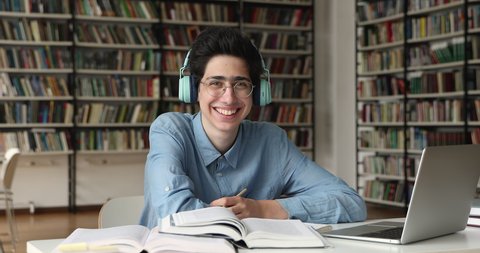 Student guy in eyeglasses and headphones sit at table with laptop and heap of textbooks, studying alone in college library, smile looking at camera. Education, e-learning, gen Z, modern tech concept