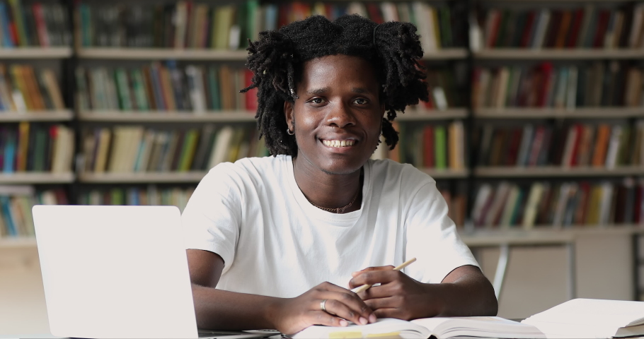 African student guy sit at table with wireless computer smile look at camera showing thumbs up hand gesture, bookshelves on background. Effective e-learn, education use modern tech, admission concept Royalty-Free Stock Footage #1083066154
