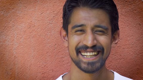 A close up portrait shot of a young handsome millennial 20 something latino man smiling with facial hair against an orange wall.