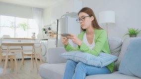 Asian frustrated beautiful woman play mobile game on cellphone at home. Attractive casual e-sport gamer girl loser sit on couch in living room, feel angry and offended due to defeat in the video game.