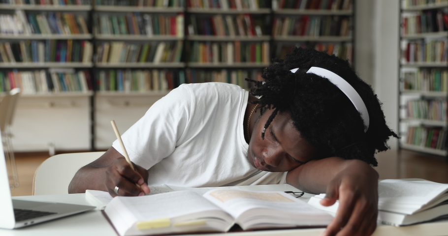 African guy student in glasses and headphones feel unmotivated prepare for college exams, makes assignment lying down on table in library looks disinterested to learn. Fatigue, bored to study concept Royalty-Free Stock Footage #1083071704