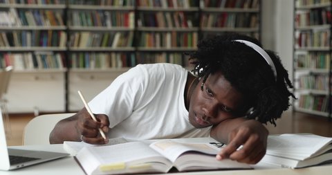 African guy student in glasses and headphones feel unmotivated prepare for college exams, makes assignment lying down on table in library looks disinterested to learn. Fatigue, bored to study concept
