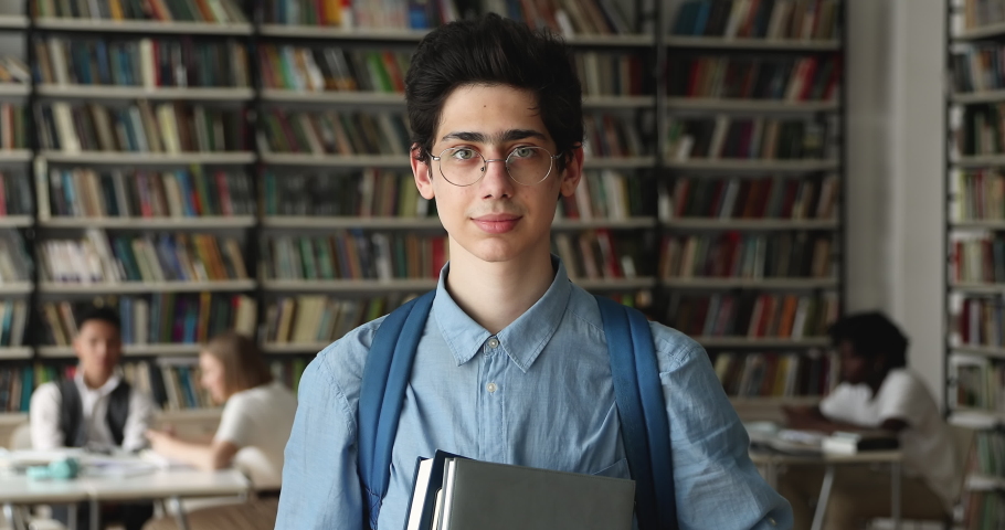 Smiling 16s high school guy schoolboy with backpack holding textbooks posing in university library. Head shot of higher education institution student portrait, excellent studies, studentship concept Royalty-Free Stock Footage #1083071755