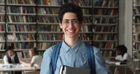 Smiling 16s high school guy schoolboy with backpack holding textbooks posing in university library. Head shot of higher education institution student portrait, excellent studies, studentship concept