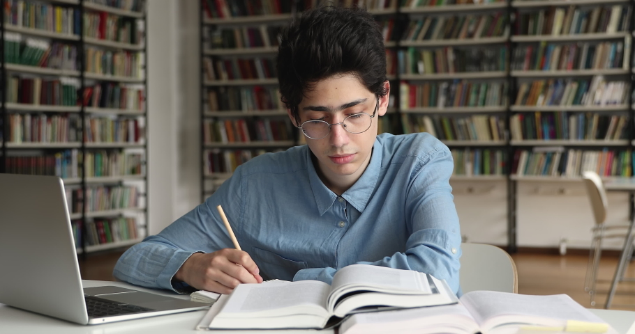 High school student in eyeglasses sit at table with computer and heap of textbooks in library read topics writing in copybook, do school work looks focused. Education, new knowledge, studying concept Royalty-Free Stock Footage #1083071770