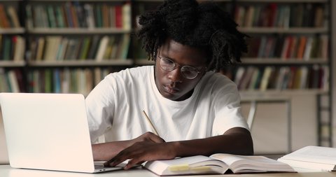 African student in glasses sit in library take notes studying with laptop, make assignment, use internet resource website, e-learn busy in self-education. Prepare for exams, gen Z, modern tech concept
