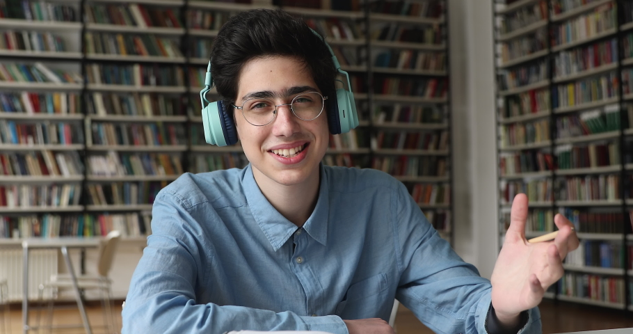 Smiling teenager guy wear wireless headphones sit at table in college library talks to camera, take part in seminar, make video call. Tuition, generation Z and on-line studies, virtual meeting concept Royalty-Free Stock Footage #1083072055