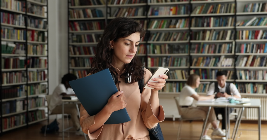 Attractive student girl standing in library holds cell phone, texting message, chatting in social media, check on-line studies timetable using mobile application, gen Z and modern tech usage concept | Shutterstock HD Video #1083072076