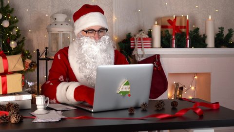 Santa Claus reading a letter and types on laptop computer at Christmas workshop.