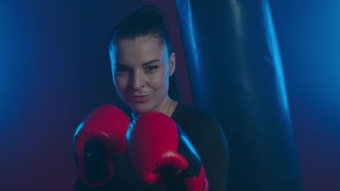 Female fighter in boxing gloves with an ironic smile looks at her enemy and challenges him to battle. Strong girl boxer in the ring is ready to fight and calls her opponent to start looking at camera