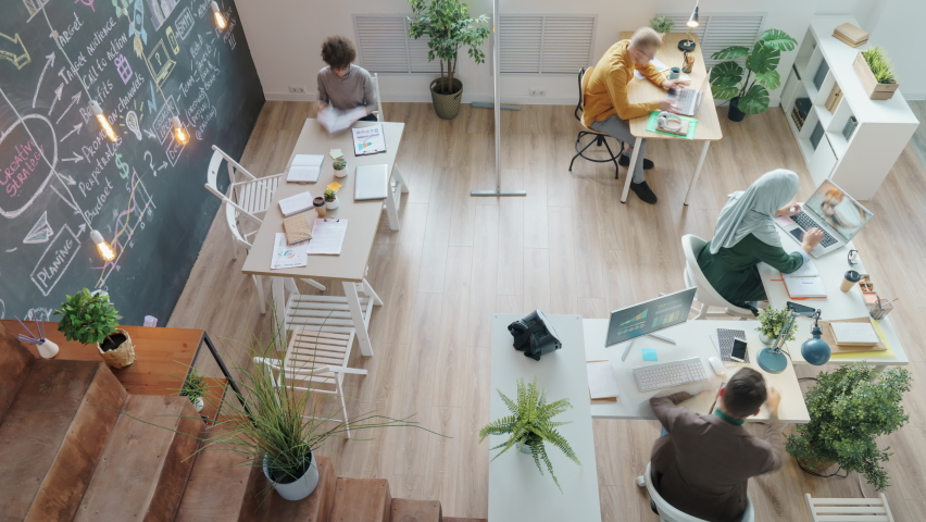 Zoom-in high angle view of diverse creative team working in shared office talking cooperating using laptops writing on chalkboard wall | Shutterstock HD Video #1083074923