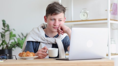 Online home school for teenage students. Handsome brunet kid drinks tea while listens spbi teacher explaining via laptop at table in room close view 4k video
