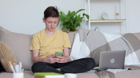 Teenage boy home online schooling. Senior schoolboy in casual t-shirt reads message spbi on mobile phone sitting near laptop computer on couch in living room 4k video