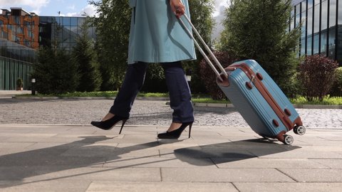 Closeup view of young woman walking through city with suitcase in hand and heading to airport spbi. Legs pic of beautiful businesswoman holds luggage and strolls confidently along glass building in