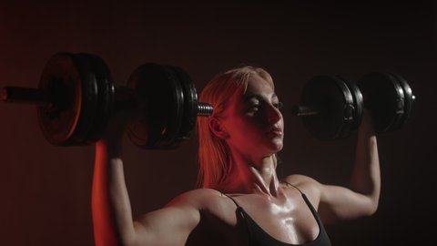 A fitness model in a bodysuit is doing a dumbbell press over his head. She is performing a heavy set on her shoulder. An orange light is shining on her. 4K 50fps