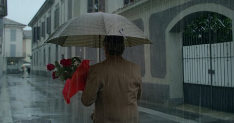 Cinematic shot of romantic happy senior pensioner man with umbrella and bouquet of red roses walking in city center under rain to bring them to his beloved woman.