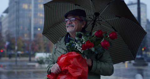 Cinematic shot of romantic happy senior pensioner man with umbrella and bouquet of red roses walking in busy city center under rain to bring them to his beloved woman.