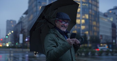 Cinematic shot of smiling senior pensioner man with umbrella is using smartphone for send messages his family or navigate in internet while walking in city center under rain in evening.