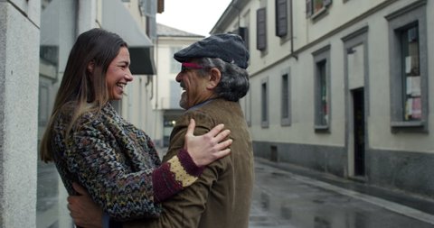Cinematic shot of elderly father makes surprise visit to his daughter at home, she is happy to see he and giving affective hug as sign of love and respect in city center under rain.