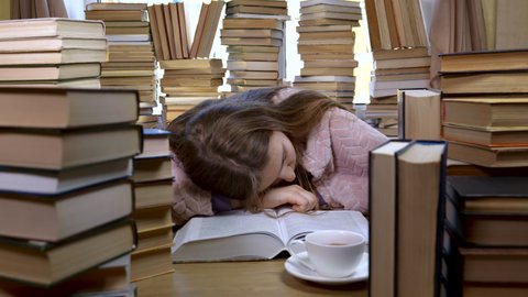 A girl reads a book in the library. Stacks of books and cup of tea on table. The girl is tired of studying and wants to sleep. Concept of Reading and Education. Ultra 4K