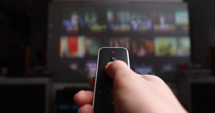 Womans hand selects internet tv channels with remote control. Close up view. Blurry tv at the background Royalty-Free Stock Footage #1083081613