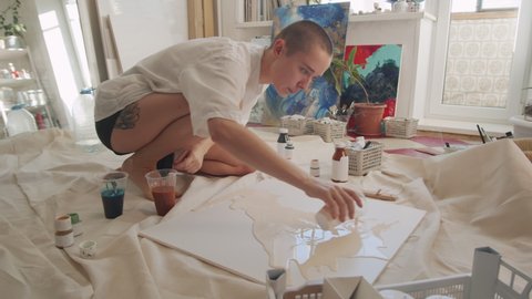 Artist wearing white shirt sitting on floor in studio starting new artwork pouring acrylic paint on canvas
