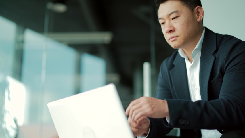 Portrait thoughtful Asian businessman working on a laptop computer at a modern office desk. Confident Focused pensive man in formal suit indoors. thinking of inspiration solving a problem. Startup Royalty-Free Stock Footage #1083084748