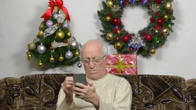 Against the background of a Christmas wreath and a Christmas tree, an old man looks at the phone