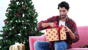 4k video of A handsome caucasian young man happily hugging a gift box sitting on the sofa in a room decorated with Christmas trees and festive atmosphere.