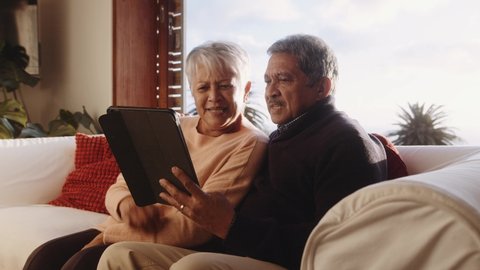Multi-cultural elderly couple sitting on sofa, laughing at tablet. High quality 4k footage