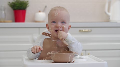 Closeup of young baby in white feeding high chair, kid is trying to eat himself, happy child with food stained face, little boy eats porridge with a spoon. High quality 4k footage Video de stock