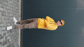 Young Man caucasian dancing moving his hands with his legs with making funny faces wearing yellow hoodie, beanie hat sunglasses close up against blue sky. Younger generation. Lifestyle. Vertical video