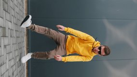 Active Young Man caucasian dancing moving his hands with his legs with making funny faces Vertical video storytelling for social media wearing yellow hoodie sunglasses close up grey wall street. Young