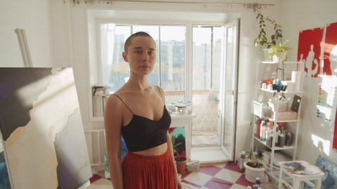 Zoom in shot footage of stylish young woman wearing crop top and skirt standing in studio full of her paintings looking at camera