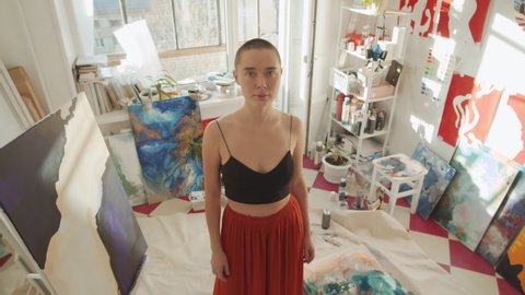 High angle view shot of beautiful young woman wearing crop top and skirt standing in studio full of her artworks looking at camera