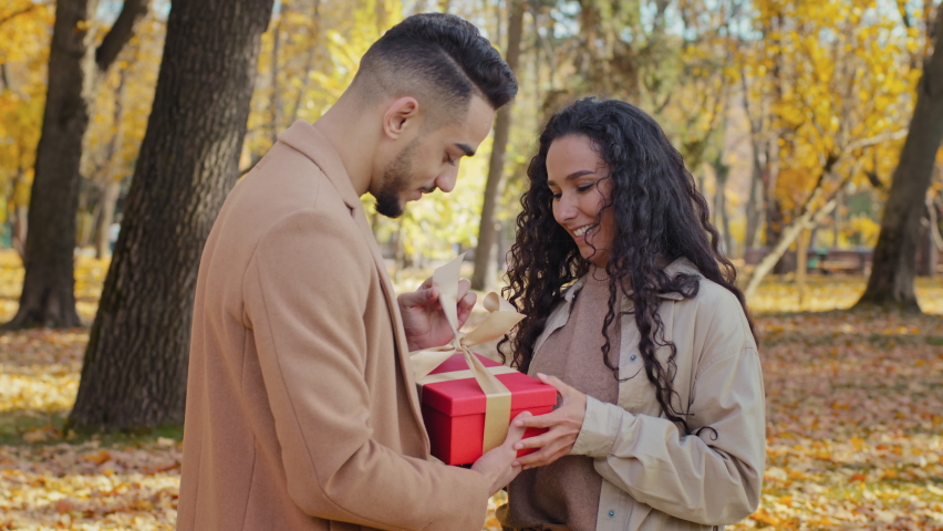Girl gives gift to beloved boyfriend young guy opens red box unleashing gold ribbon smiling hispanic couple stands in autumn park bearded man surprised excited looking present family laughing have fun Royalty-Free Stock Footage #1083104899