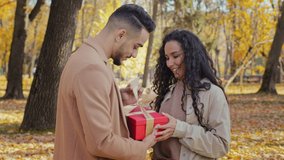 Girl gives gift to beloved boyfriend young guy opens red box unleashing gold ribbon smiling hispanic couple stands in autumn park bearded man surprised excited looking present family laughing have fun - Powered by Shutterstock - Get 15% off with code: PIKWIZARD15